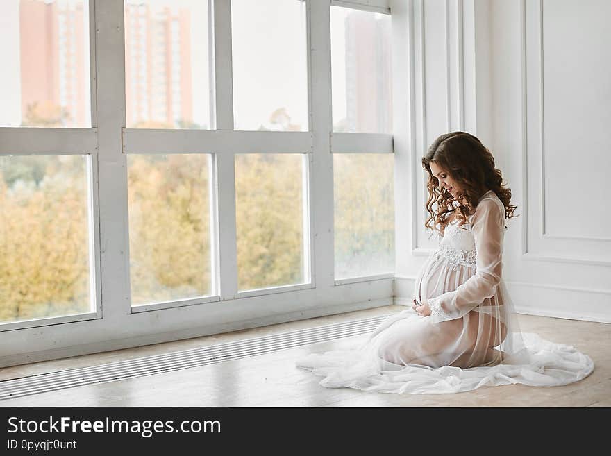 Beautiful young pregnant woman with fashionable hairstyle in a stylish peignoir smiling and posing near the big window at light interior.