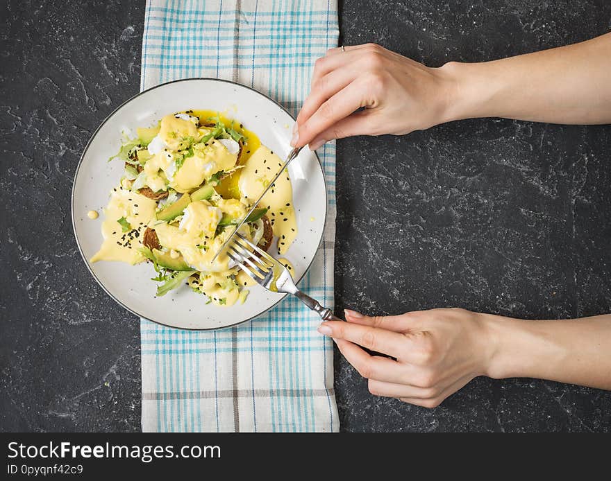 Female hand hold fork and knife. Eggs Benedict with salad and avocado on stone background. Top view