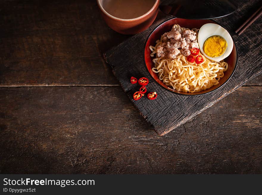 Instant noodle with pork, egg and vegetables on black bowl on the wood table there are chilli, chopstick and drinking glass placed around close up