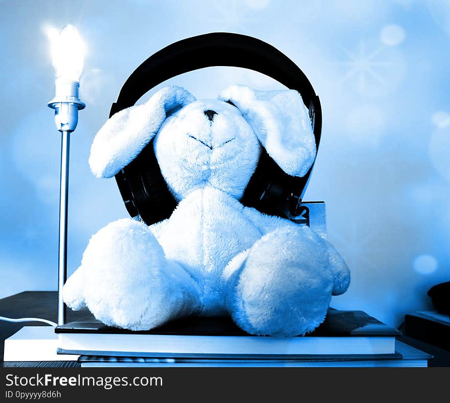 A soft rabbit plush toy with too big headphones sitting laid back on a pile of books enjoying music/audio-book. A lit table lamp stands next to it. Soft blue bokeh effect layer. A soft rabbit plush toy with too big headphones sitting laid back on a pile of books enjoying music/audio-book. A lit table lamp stands next to it. Soft blue bokeh effect layer.