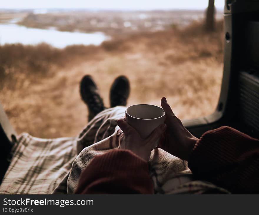 Women legs in leather shoes wrapped in a plaid lie in the trunk of a car with an open door, overlooking a rural landscape.