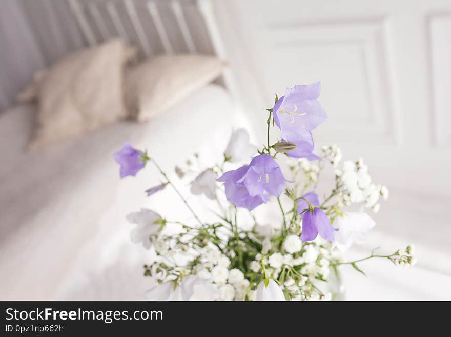 Glass vase with lilac and white floweers  in light cozy bedroom interior. White wall, bed with white linen, light blanket or plaid and pillows