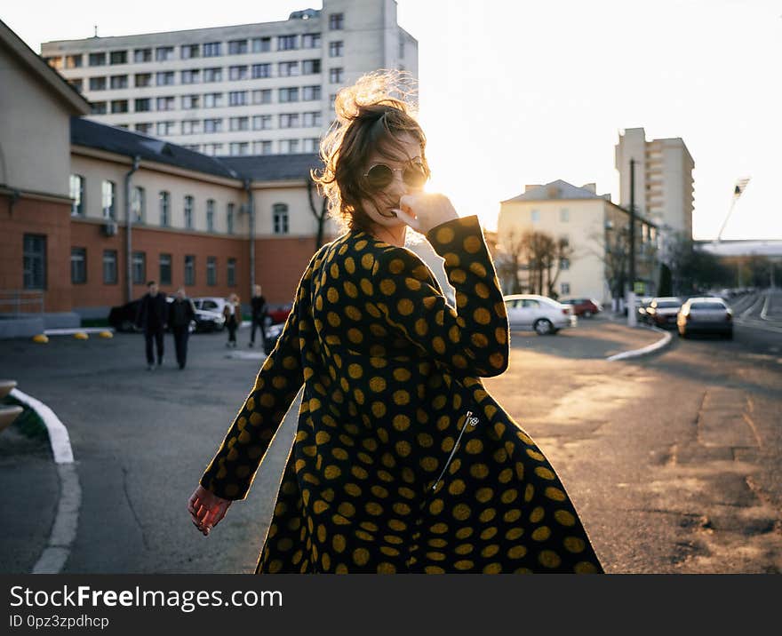 Attractive young woman with wind disheveled hair wearing polka dot coat and fashion sunglasses turns around at camera on street at sunset. Attractive young woman with wind disheveled hair wearing polka dot coat and fashion sunglasses turns around at camera on street at sunset