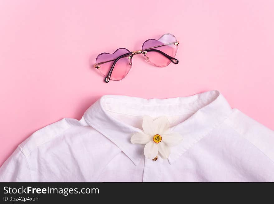 Collarless white shirt with stylish heart-shaped glasses and a spring flower on a pink background. Top view, flat lay. Collarless white shirt with stylish heart-shaped glasses and a spring flower on a pink background. Top view, flat lay