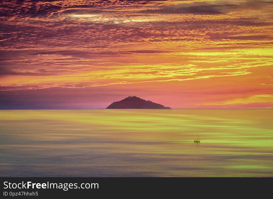 A fiery sunset with a boat that travels in front of an island and a sky that is reflected in the sea