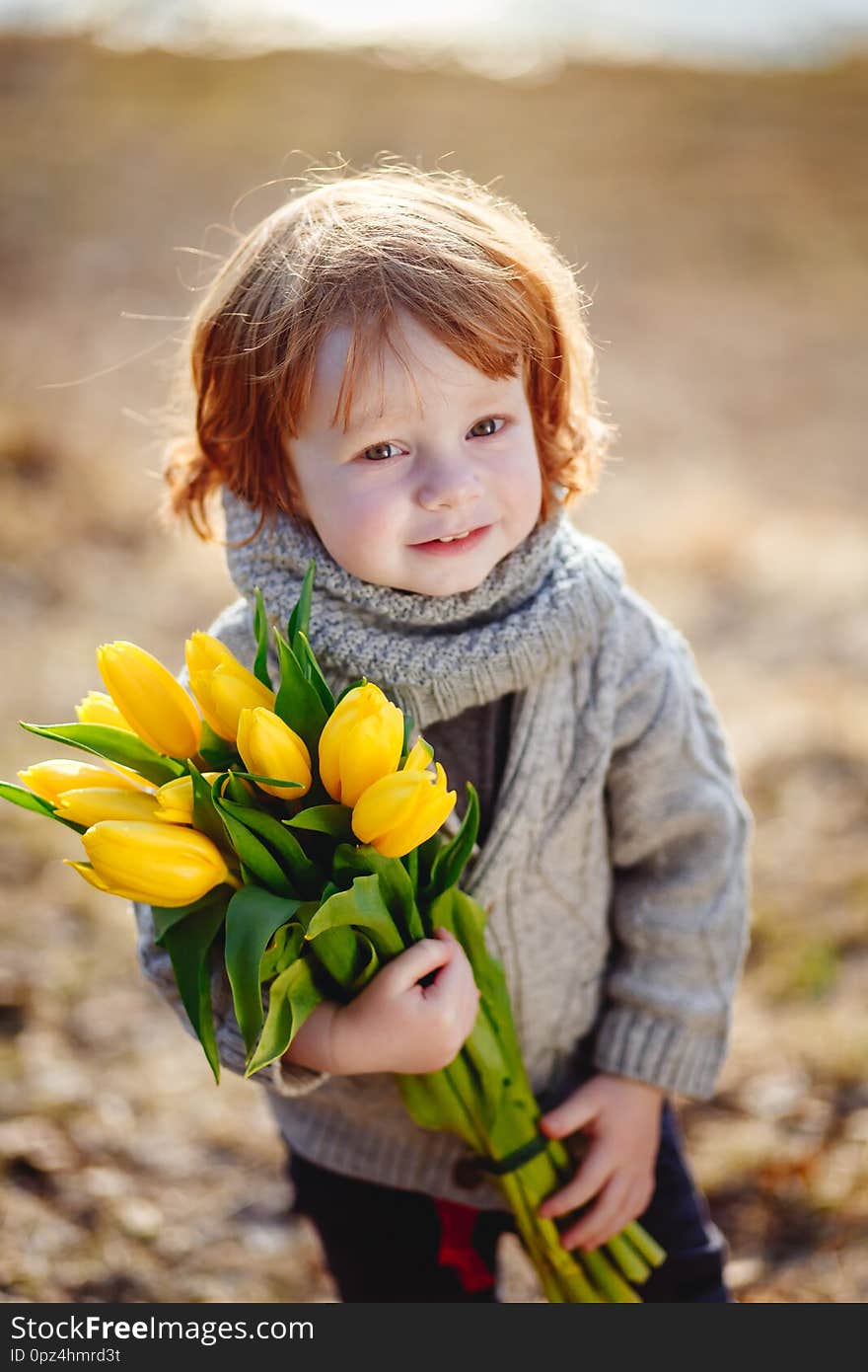 A cheerful cute little boy with red hair holding yellow tulips outside in early spring in the forest near the water.