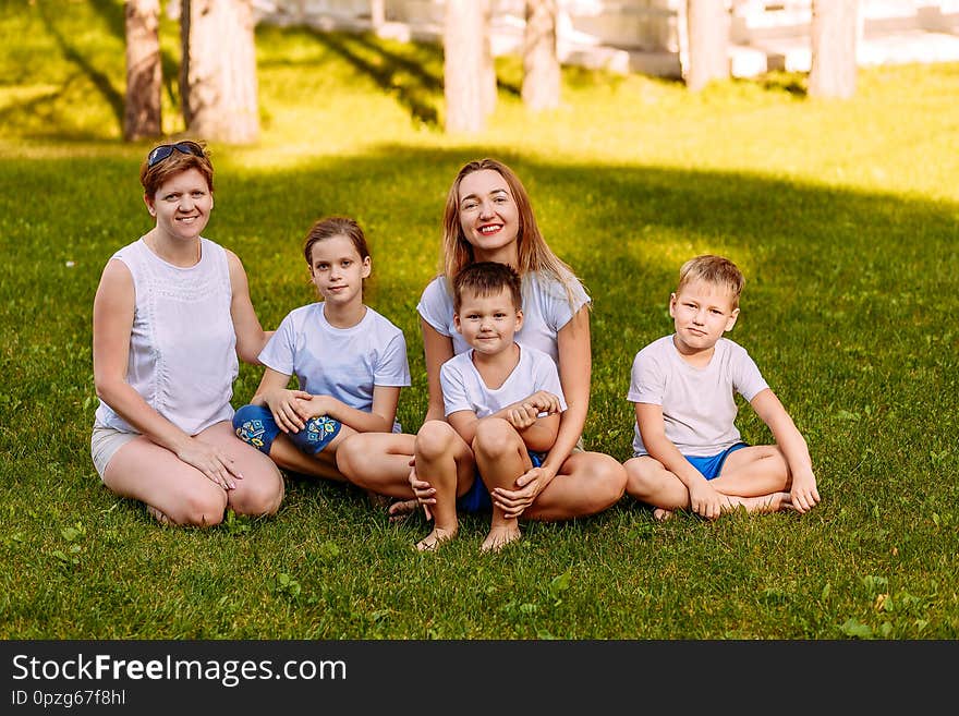 Happy women and children sit on the green grass and look at the camera. Big happy family, two mothers and three children in white