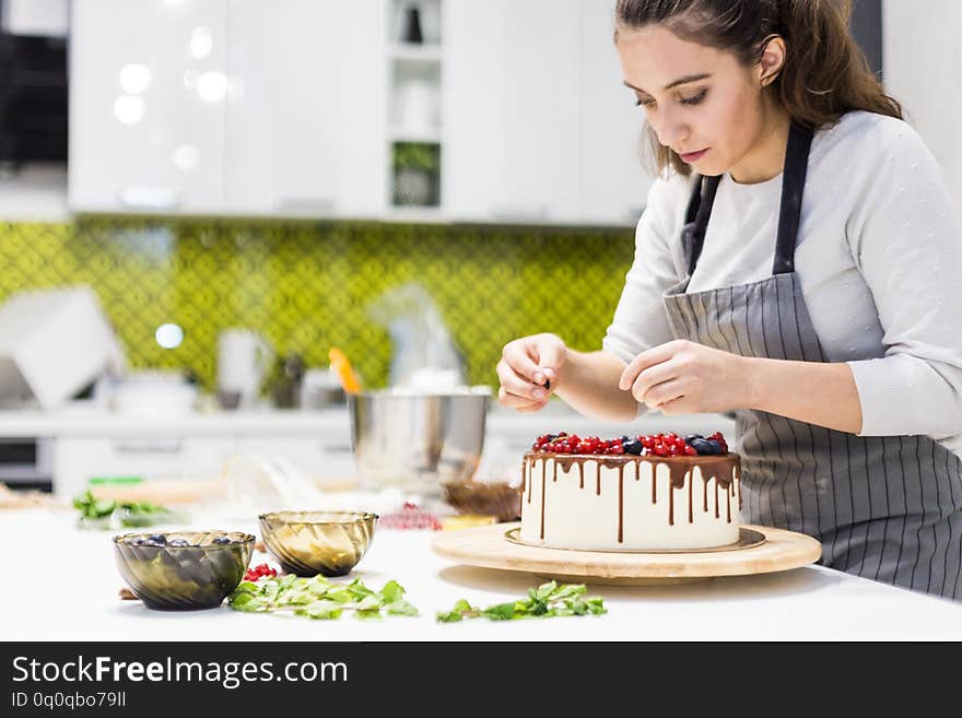Confectioner decorates with berries a biscuit cake with white cream and chocolate. Cake stands on a wooden stand on a white table. The concept of homemade pastry, cooking cakes