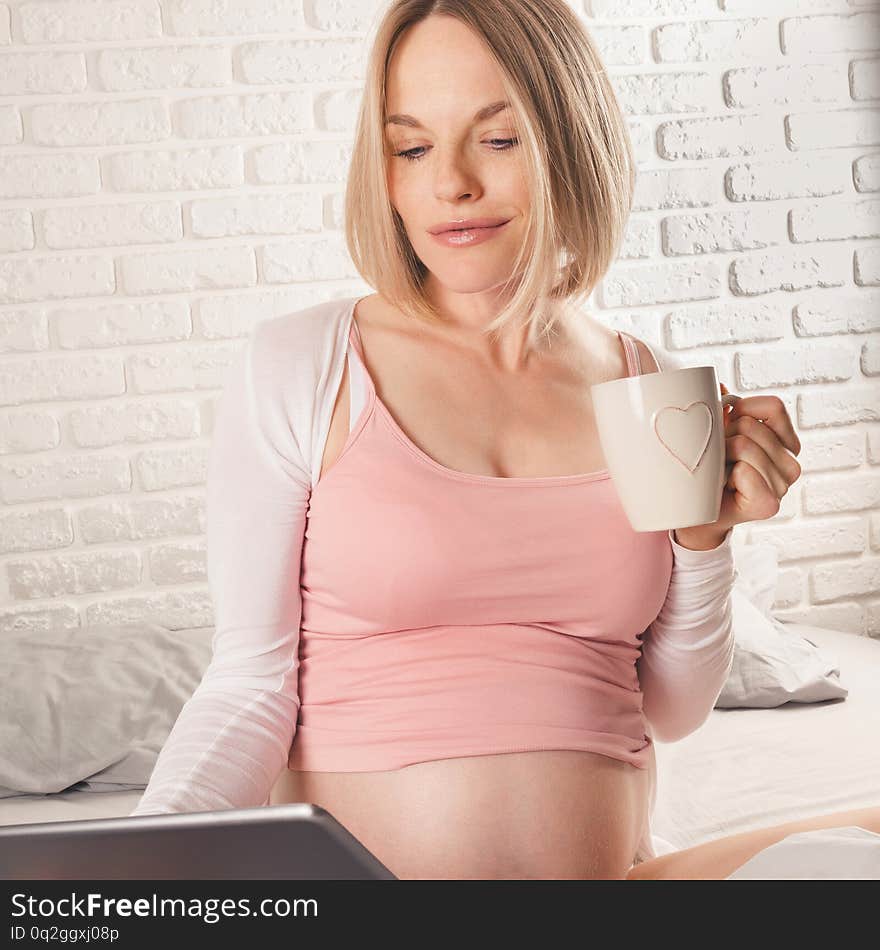 Happy smiling pregnant woman is sitting in bed and looks at tablet.