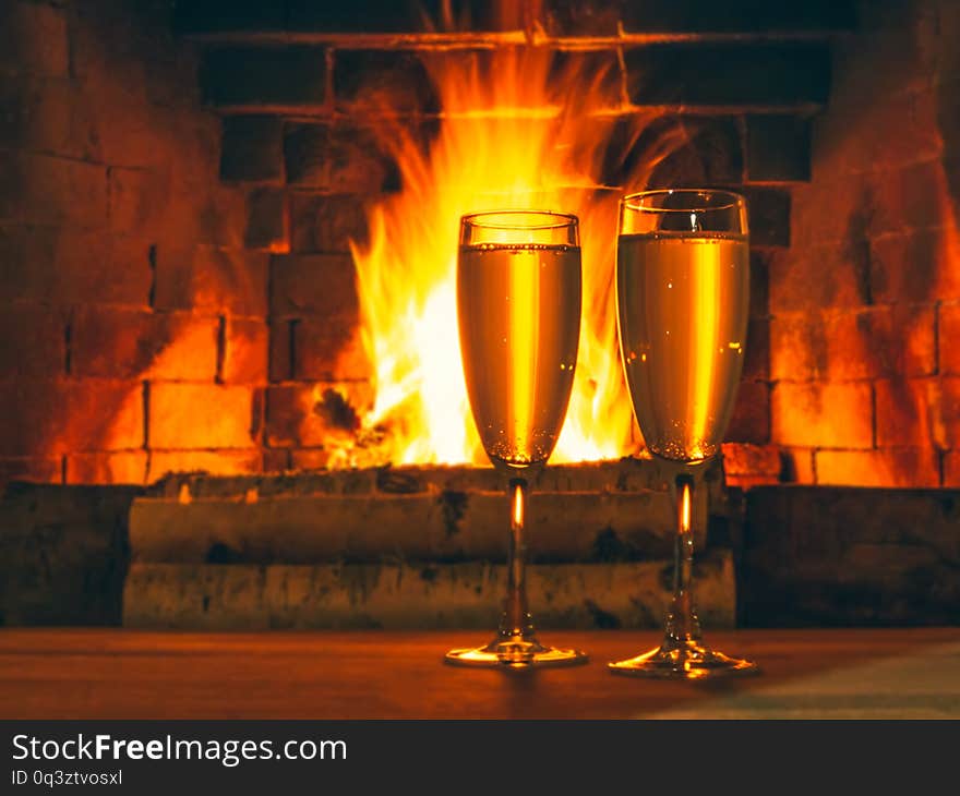 Two glasses with champagne on a wooden table near the fireplace with burning birch wood.