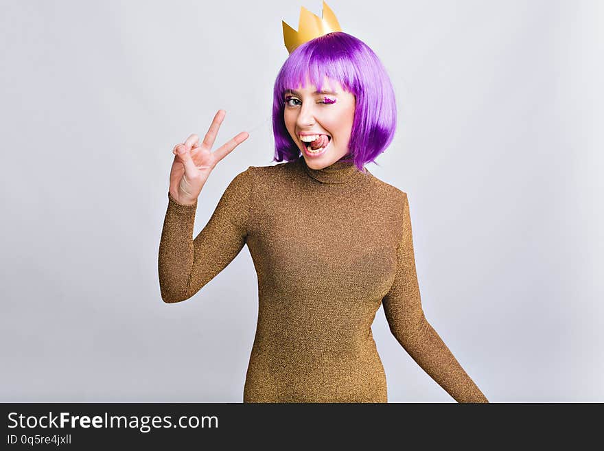 Portrait pretty girl with gold crown having fun in studio. She wears violet haircut, shows tongue and looks happy to