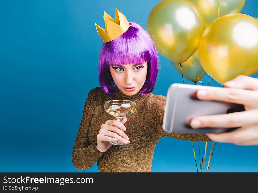 Charming young woman with purple haircut, crown on head making selfie portrait on blue background. Golden balloons, champagne, celebrating new year party, luxury dress, tinsels makeup