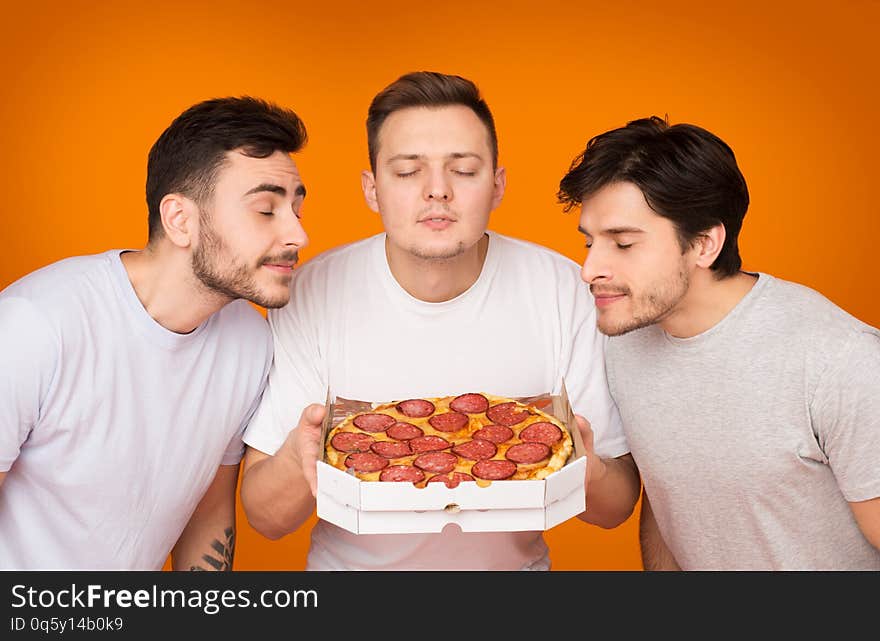 Three Men Smelling Hot Pizza Over Orange Studio Background