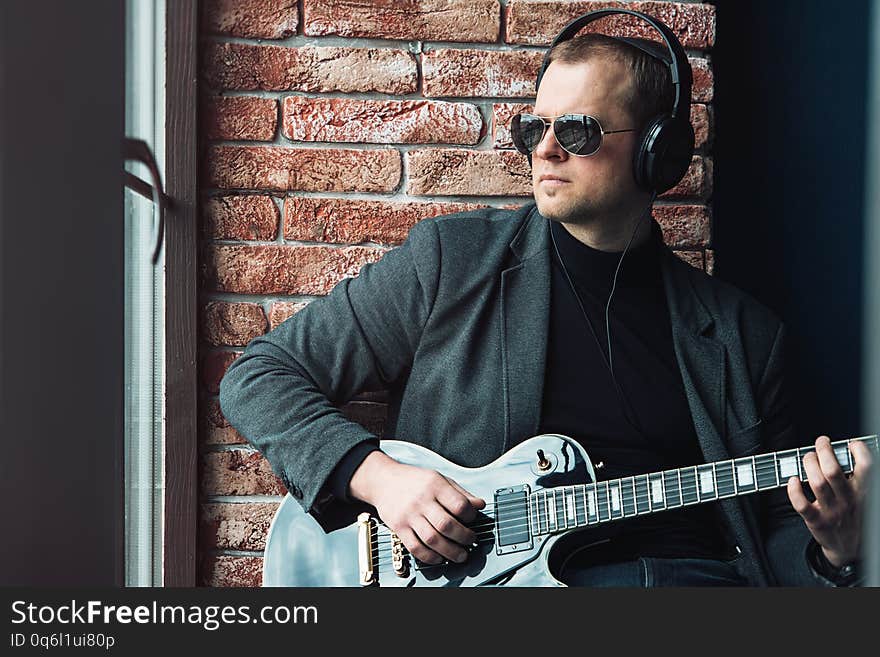 Man singer sitting on a window sill in a headphones with a guitar recording a track in a home studio. Man wearing sunglasses, jeans, black shirt and a jacket