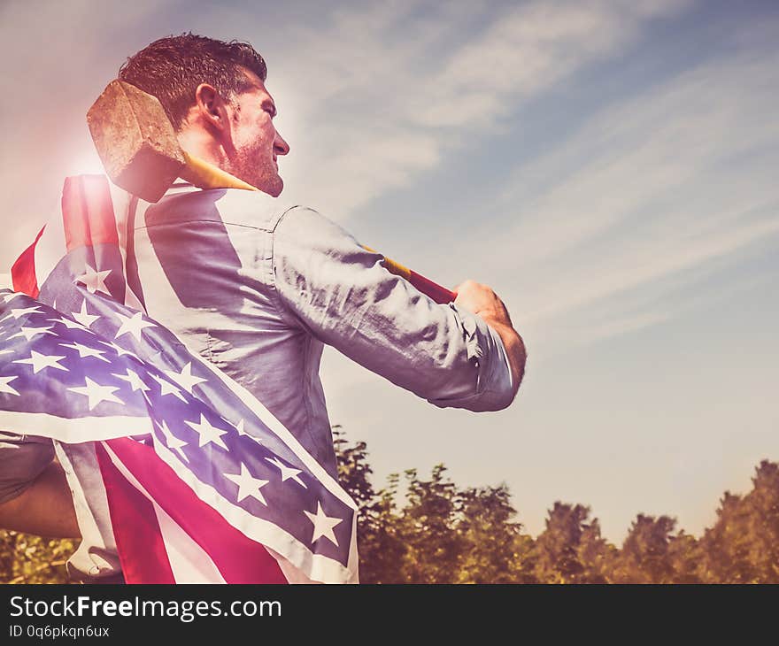 Attractive man holding a sledgehammer and a US Flag in his hands and looking into the distance against a background of trees, blue sky and the rays of the setting sun. National holiday concept. Attractive man holding a sledgehammer and a US Flag in his hands and looking into the distance against a background of trees, blue sky and the rays of the setting sun. National holiday concept