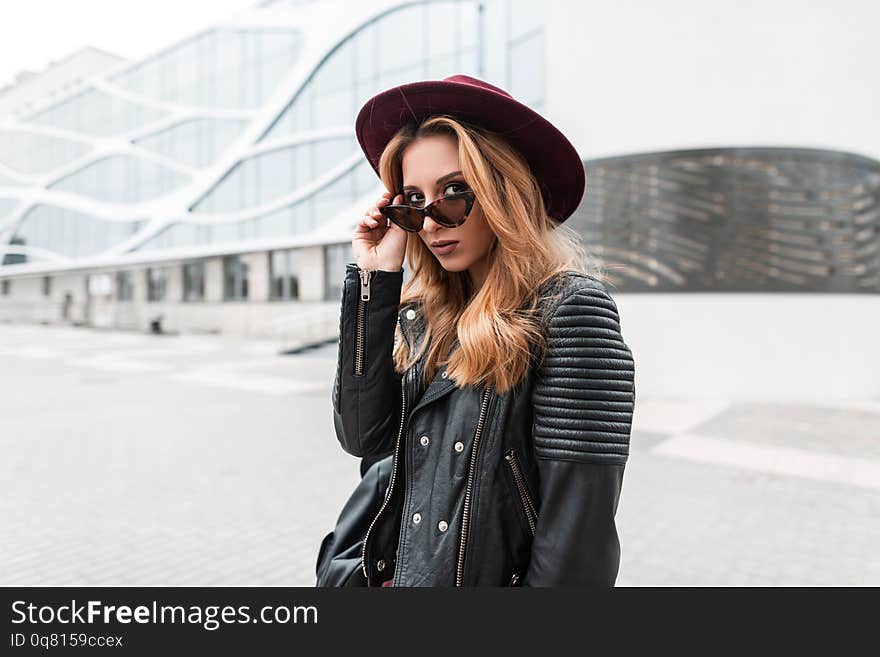 Sexy pretty young hipster woman in sunglasses in vintage hat in trendy leather jacket with black backpack stands and looking in the camera outdoors. Urban girl walks through the city. Youth style