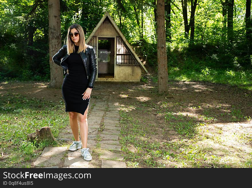 Beautiful fashion model girl with sunglasses, black dress and leather jacket in nature