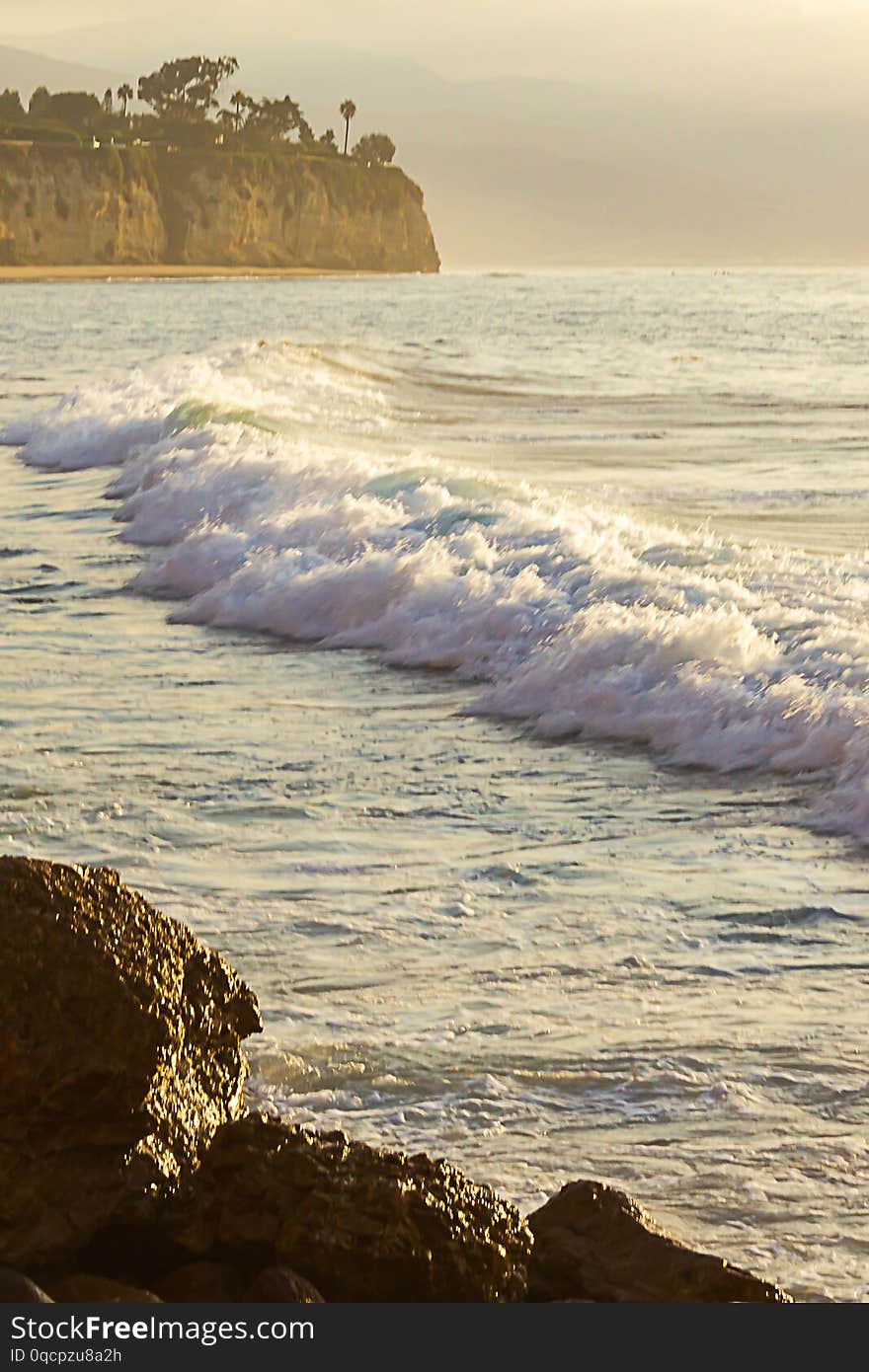 Foaming wave breaking onto ocean espanse with cliff and boulders at sunrise