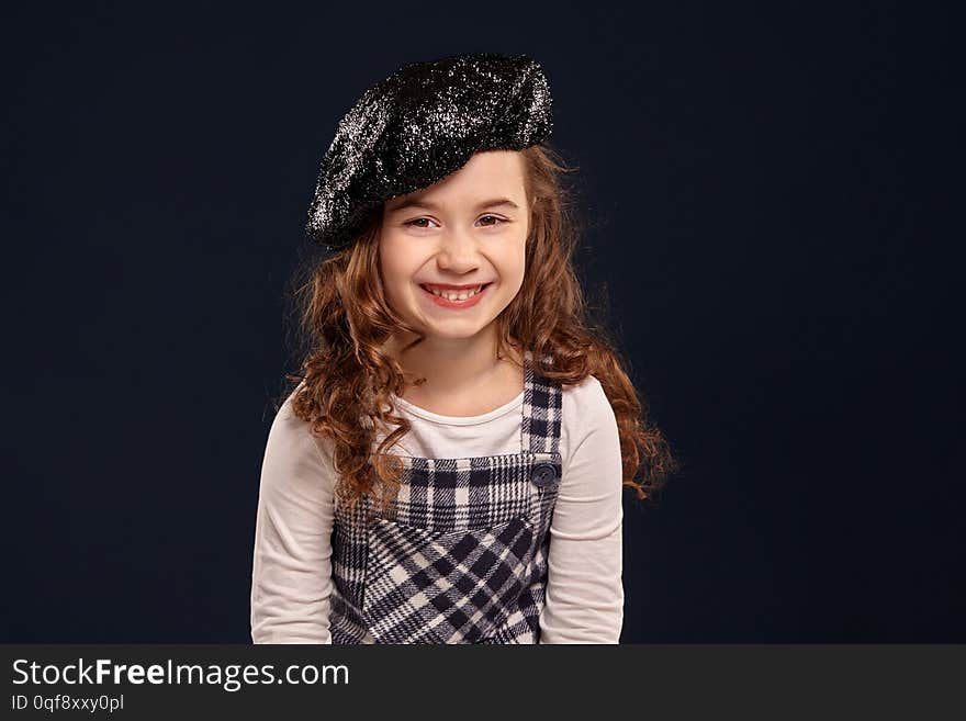 Pretty little child with a beautiful curly hair wearing checkered dress, white blouse and a black sparkling beret is smiling widely and looking at the camera. Stylish brunette kid is posing in studio on a black background. Children`s fashion.