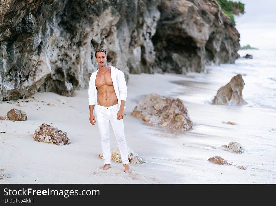 Sexy tanned man in white shirt, pants rests on beach, ocean waves at background