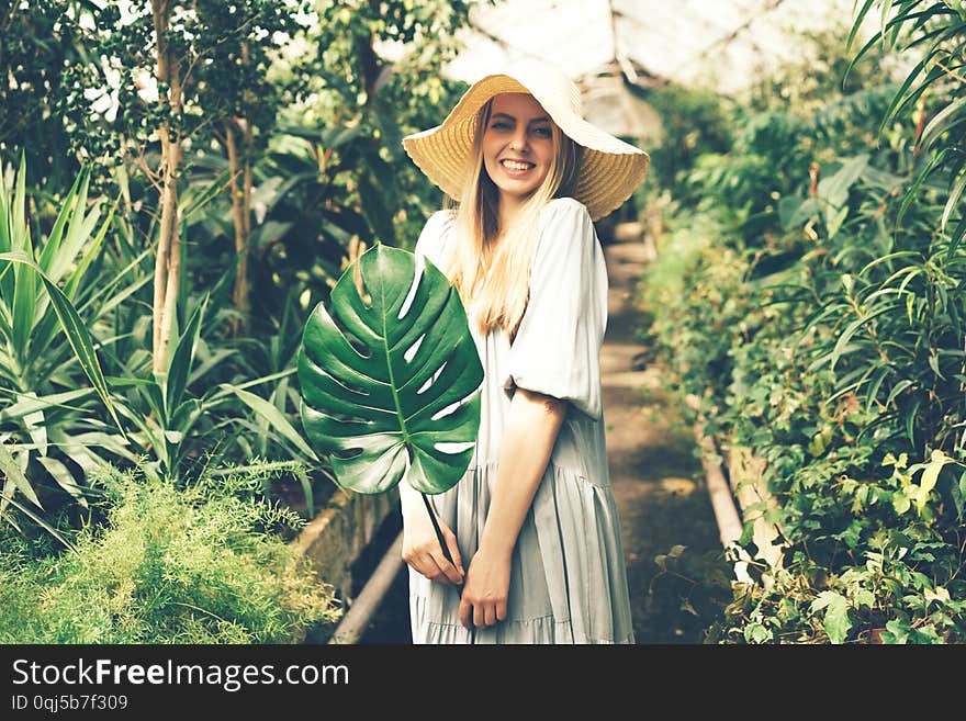 Young beautiful blonde in a dress and straw hat in a tropical greenhouse with monstera leaf in her hands, happy vacation days, summer mood concept. Young beautiful blonde in a dress and straw hat in a tropical greenhouse with monstera leaf in her hands, happy vacation days, summer mood concept