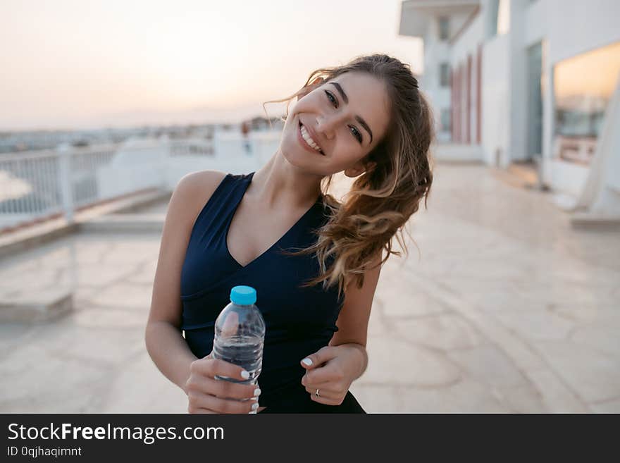 Portrait charming attractive young woman in sportswear with bottle of water at training in sunny morning on seafront in tropical country. Outwork, smiling to camera, expressing positivity