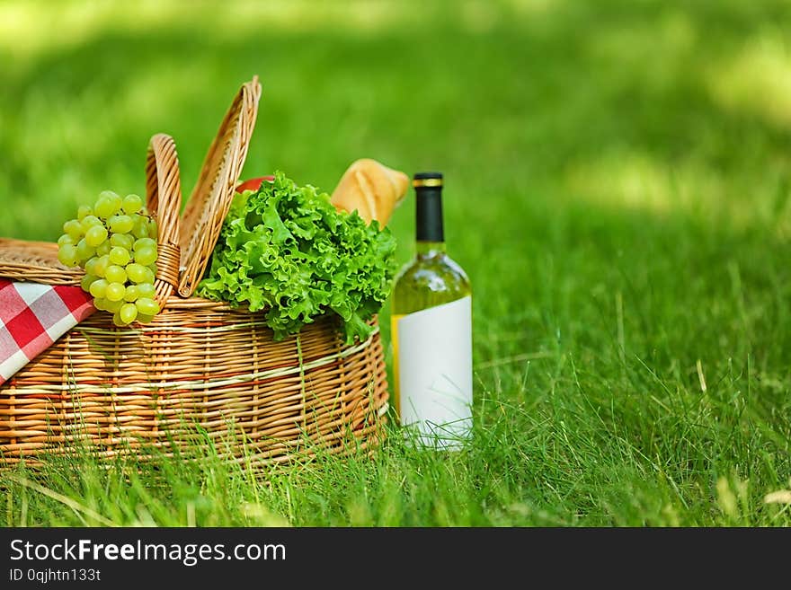 Wicker basket with blanket, wine and food on green grass in park, space for text. Summer picnic. Wicker basket with blanket, wine and food on green grass in park, space for text. Summer picnic