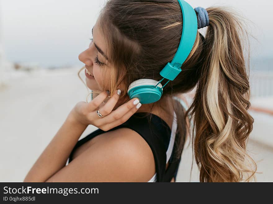 Closeup portrait joyful amazing woman in sportswear, with long curly hair listening to music through blue headphones, walking on seafront. Cheerful mood, fitness outside, fashionable model