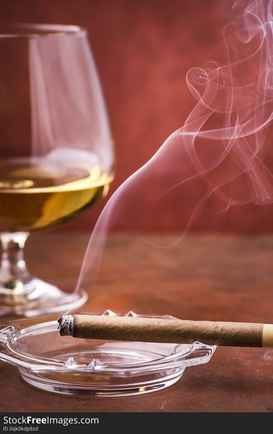 A lit cigar with smoke on ashtray in the foreground, in the background a glass with alcohol. A lit cigar with smoke on ashtray in the foreground, in the background a glass with alcohol