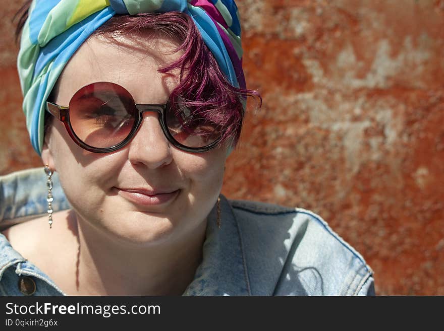 Portrait of a cheerful girl in a colored turban wearing round glasses and a denim jacket on a background of rusty iron. Portrait of a cheerful girl in a colored turban wearing round glasses and a denim jacket on a background of rusty iron