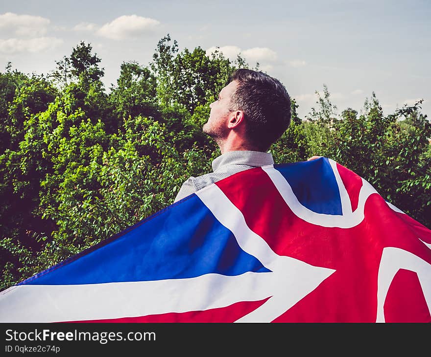 Attractive man waving a British Flag against a background of trees and blue sky. View from the back, close-up. National holiday concept. Attractive man waving a British Flag against a background of trees and blue sky. View from the back, close-up. National holiday concept