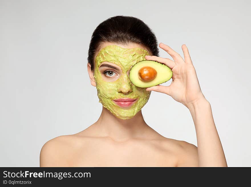 Beautiful young woman with facial mask and fresh avocado on light background