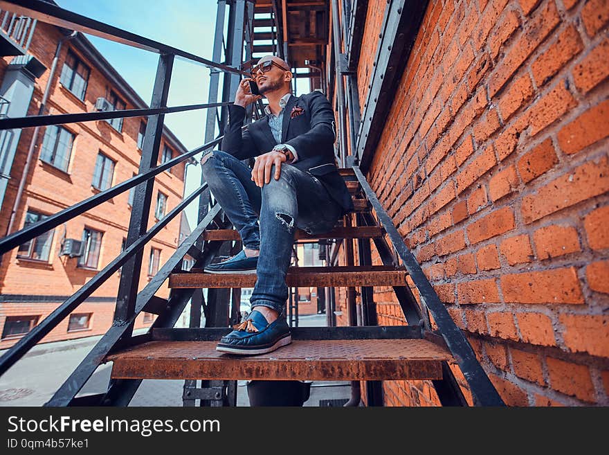 Handsome groomed man in blazer and sunglasses is talking by mobile phone while sitting on the stairs. Handsome groomed man in blazer and sunglasses is talking by mobile phone while sitting on the stairs.