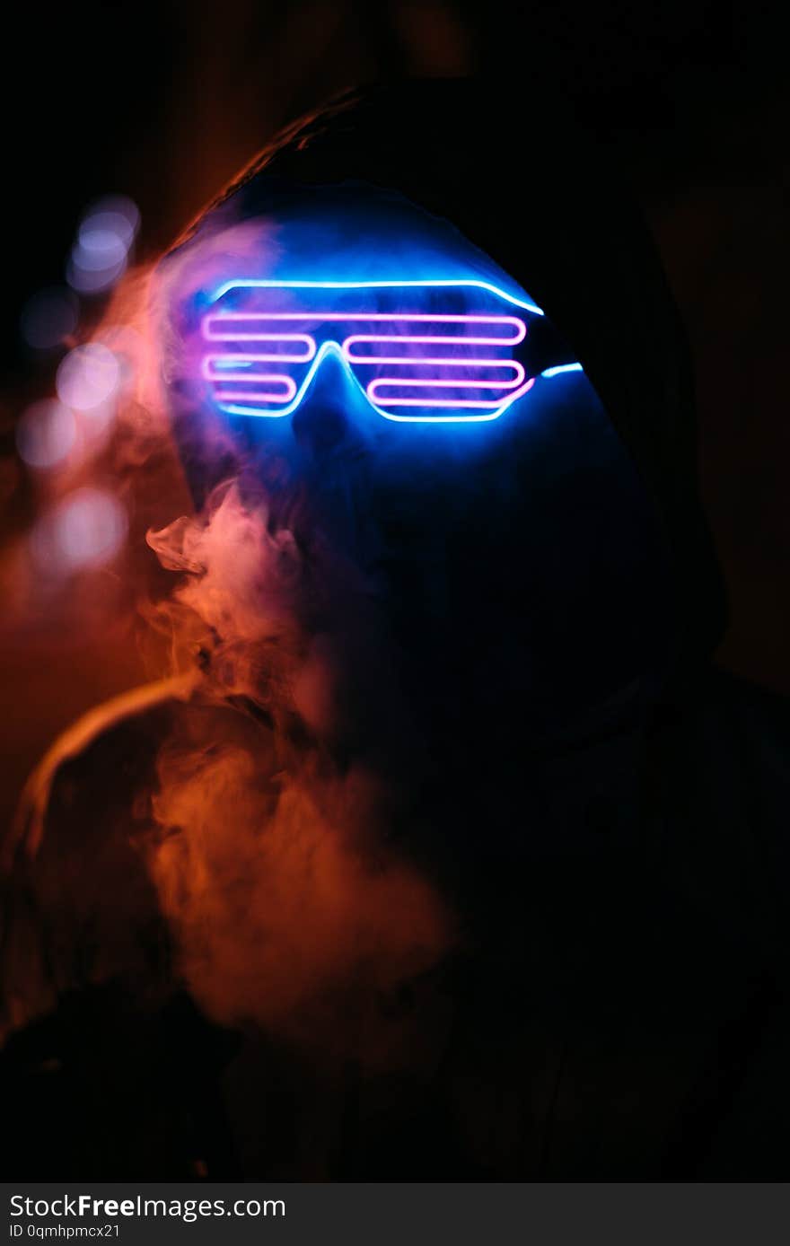 Head in neon paint and glowing glasses in a hood a man close-up in neon paint smokes, face in neon and smoke