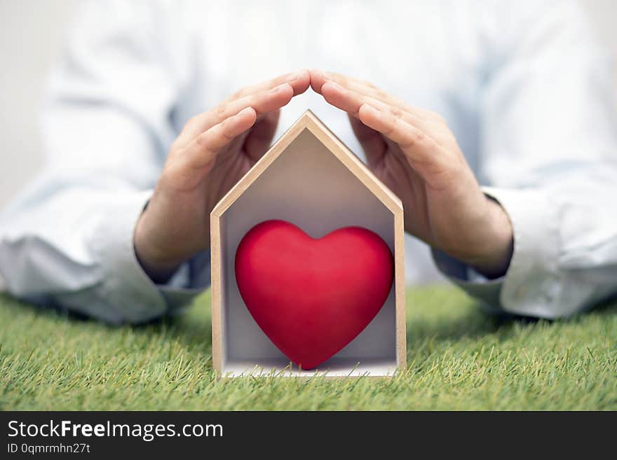 Small wooden house with red heart on green grass protected by hands