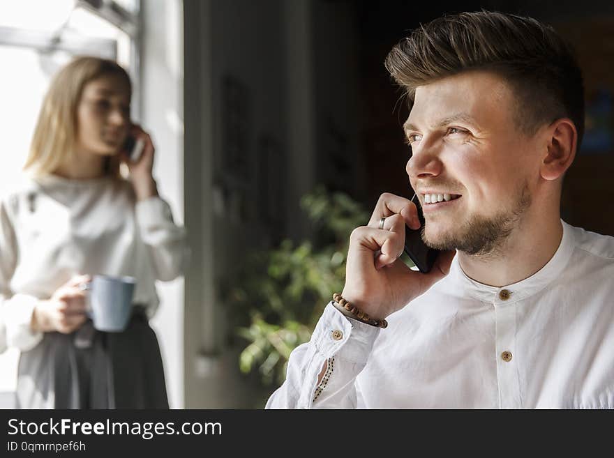 Nice and handsome man is sitting and talking on the phone. He is smiling and looking straight. Girl is standing besides window and