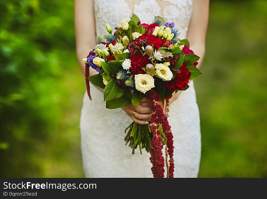 A beautiful colorful bouquet of fresh various flowers in the hands of the young beauty in wedding dress outdoors in summer day.