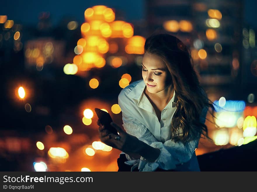 Young woman holding a smart phone in hands texting in the evening with city light on background. Young woman holding a smart phone in hands texting in the evening with city light on background