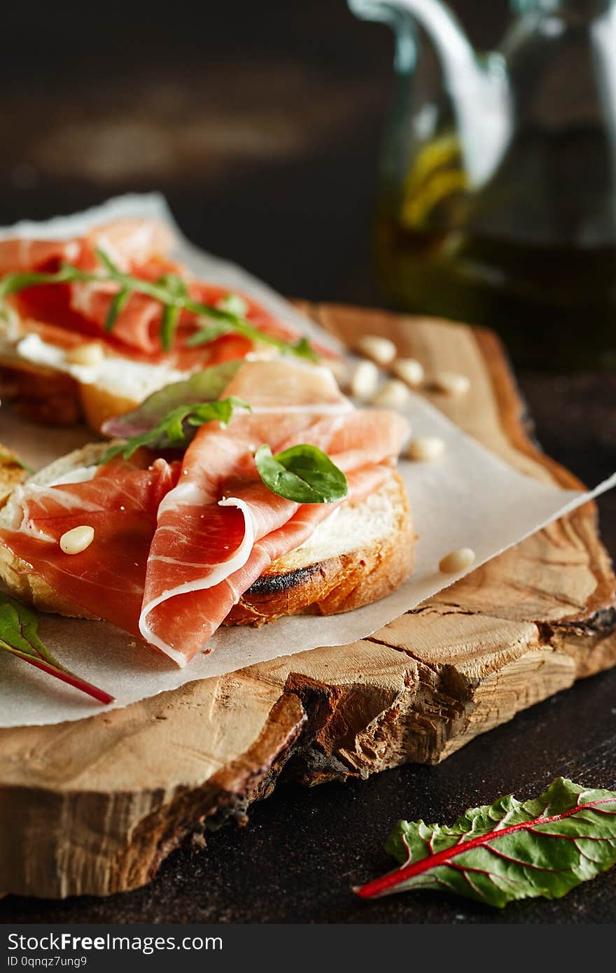 Traditional parma cured ham antipasto. Bruschetta set with Parma Ham and Parmesan Cheese. Small sandwiches with prosciutto, parmesan cheese, fresh arugula, olives and pine nuts on rustic wooden board