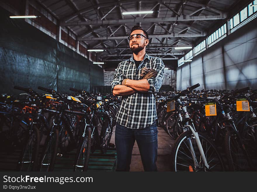 Pensive man in protective glasses and checkered shirt is posing at his own warehouse full of bicycles. Pensive man in protective glasses and checkered shirt is posing at his own warehouse full of bicycles.