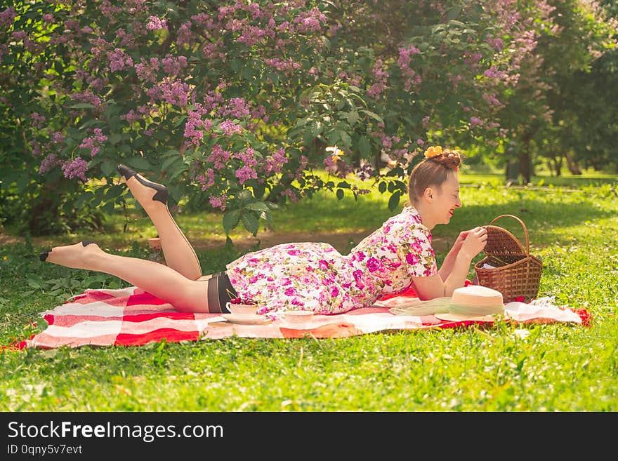 Sweetheart charming pinup girl in a summer dress on a checkered blanket in the Park near the bushes of lilac enjoys life and leisure alone. pretty happy caucasian woman on the grass. happy