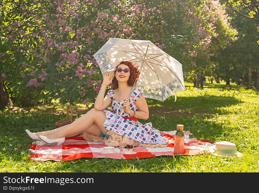 Redhaired pretty pinup woman in vintage summer dress and classic stockings with a seam in the back sits on a blanket on the grass in the city Park, next to a lilac Bush. happy girl with sun umbrella. fun