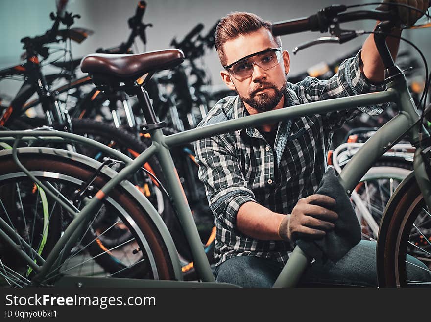 Busy attractive man in protective glasses is cleaning one of bicycle from dust.