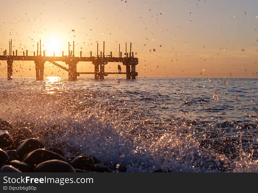 Splashing waves and blurry silhouette of couple on the pier at sunset