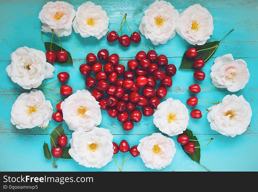 Fresh white roses with water drops, red cherry in a shape of a heart on an old painted wooden table as a bright colorful summer background for seasonal cards, posters, blogs, web design