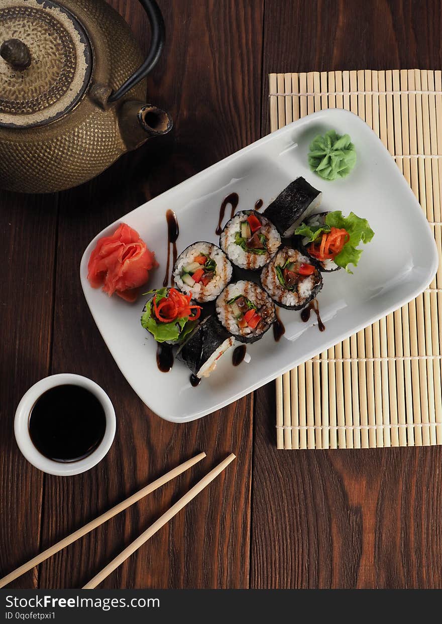 Fresh and tasty sushi on a white plate. Wooden table with a mat, chopsticks and kettle. Fresh and tasty sushi on a white plate. Wooden table with a mat, chopsticks and kettle