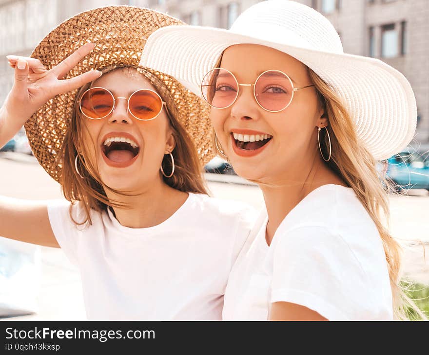 Portrait of two young beautiful blond smiling hipster girls in trendy summer white t-shirt clothes. Sexy carefree women posing on street background. Positive models having fun in sunglasses and hat. Portrait of two young beautiful blond smiling hipster girls in trendy summer white t-shirt clothes. Sexy carefree women posing on street background. Positive models having fun in sunglasses and hat