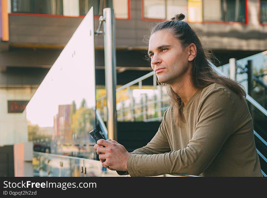 Attractive young man hipster writes a message on a smartphone while sitting on the steps of a glass business center