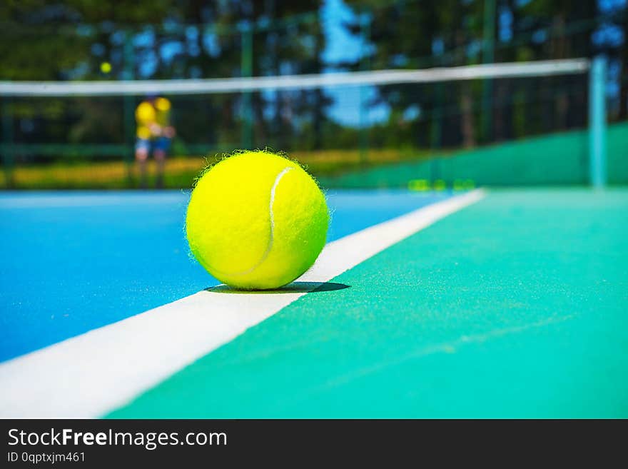 Tennis ball on white court line on hard modern blue green court with player, net, balls, trees on the background. Close-up, selective focus. Sport, tennis play, healthy lifestyle concept.