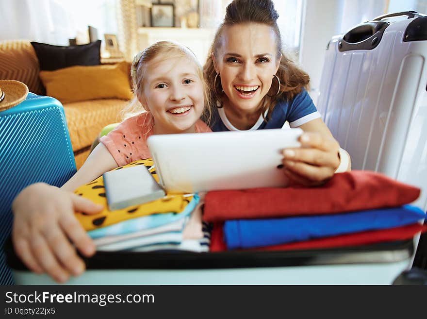 Smiling young mother and daughter tourists in the modern house in sunny summer day buying flights online using mobile app on tablet PC. Smiling young mother and daughter tourists in the modern house in sunny summer day buying flights online using mobile app on tablet PC